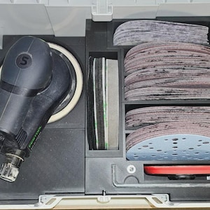 Pode incluir: Uma lixadeira orbital aleatória Festool preta com almofada de lixamento branca, guardada numa mala de plástico cinzenta com inserções de espuma. A mala contém várias pilhas de discos de lixa circulares, um prato de apoio vermelho e uma chave sextavada.