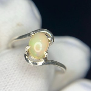 May include: A silver ring featuring a central oval-shaped cabochon opal gemstone. The metal band has a smooth, polished finish and curves around the stone, which displays iridescent flashes of light against a pale, translucent background.