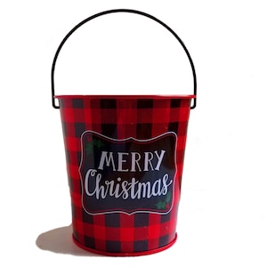 May include: Red and black plaid metal bucket with a black handle. The bucket has a white label with the text "Merry Christmas".