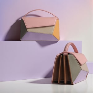 May include: Two pastel-coloured handbags with geometric patterns. The bags have a top handle and a flap closure. The colour scheme includes pink, grey, yellow, and lavender. One bag is on a purple platform, and the other is on its side.