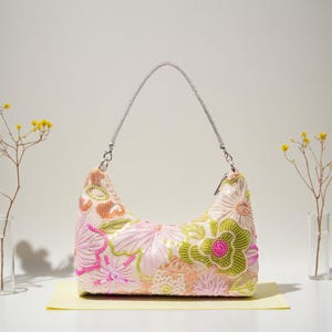 May include: A light pink handbag with a silver chain handle. The bag is decorated with embroidered flowers in shades of pink, green, and orange. The bag is on a yellow surface.