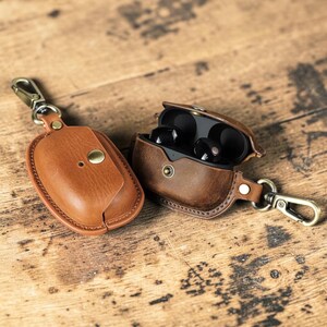 May include: Two brown leather earbud cases with gold-colored clasps and hardware. One case is closed, the other is open, revealing black earbuds. The cases are on a wooden surface.