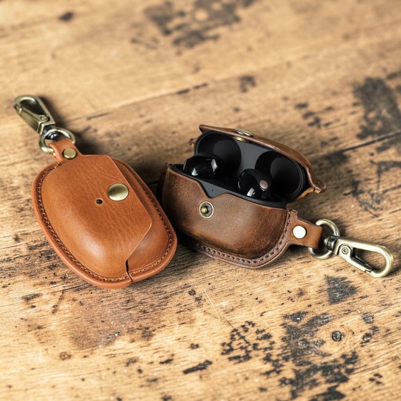 May include: Two brown leather earbud cases with gold-colored clasps and hardware. One case is closed, the other is open, revealing black earbuds. The cases are on a wooden surface.