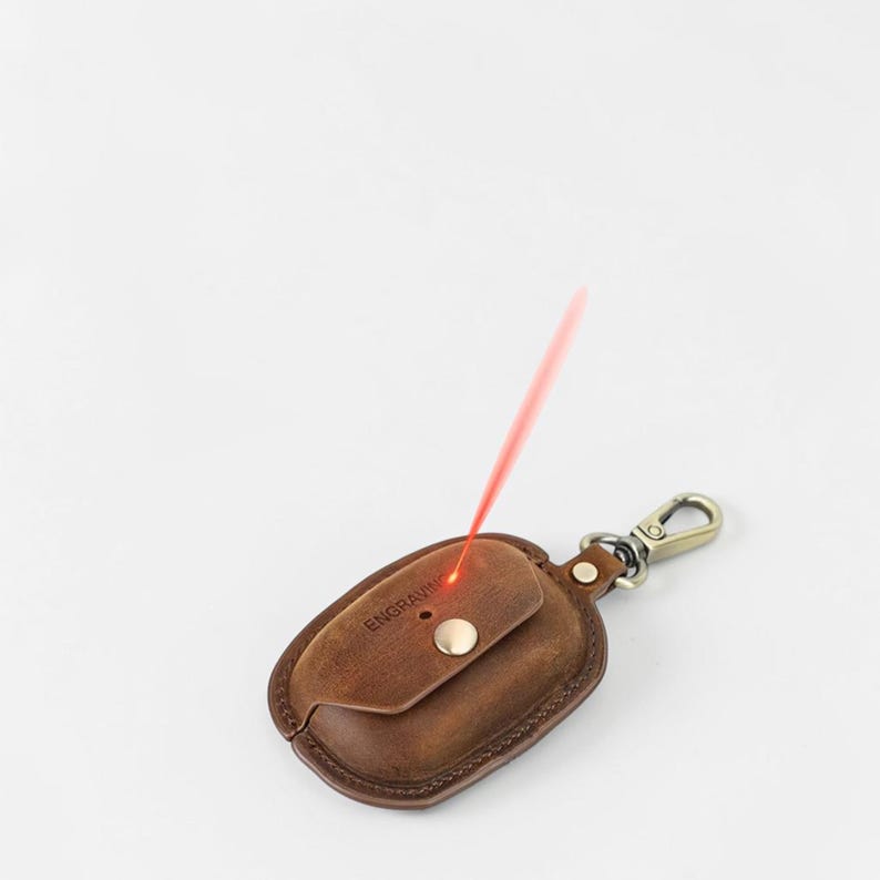 May include: A brown leather key holder with a metal clasp. The key holder has a small flap with the word "ENGRAVING" printed on it. A red laser beam is directed at the key holder.