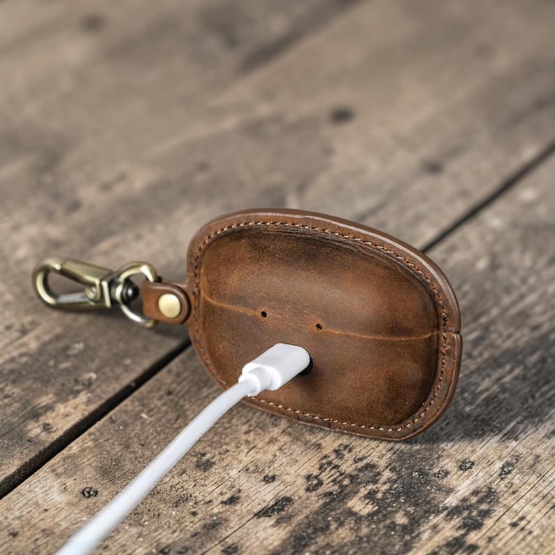 May include: A brown leather case with a gold-colored clasp and a white charging cable. The case is oval-shaped with a stitched border and two small holes. The charging cable is inserted into the case.