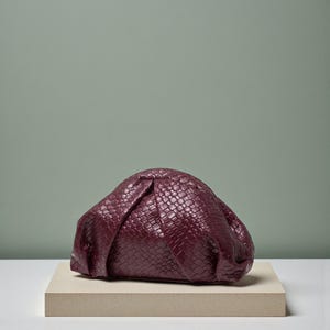 May include: A burgundy clutch bag with a woven texture, resting on a rectangular beige platform. The bag has a gathered design and a rounded shape. The background is a muted green.