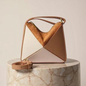 Puede incluir: Un bolso geométrico con un asa marrón y una correa para el hombro desmontable. El bolso presenta un diseño de bloques de color con paneles blancos, marrones y beige. El interior es de ante marrón claro. El bolso está sobre un pedestal de mármol.