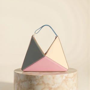 May include: A geometric handbag with a triangular design. The bag features panels of gray-green, pink, and cream-colored leather, with a blue handle. The bag is displayed on a marble pedestal against a neutral background.