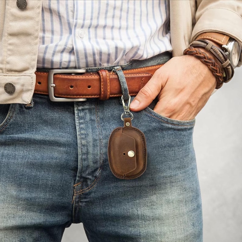 May include: A brown leather AirPod case with a snap closure is attached to a belt loop with a silver carabiner. The case is hanging from a brown leather belt with a silver buckle. The person is wearing blue jeans and a striped shirt.