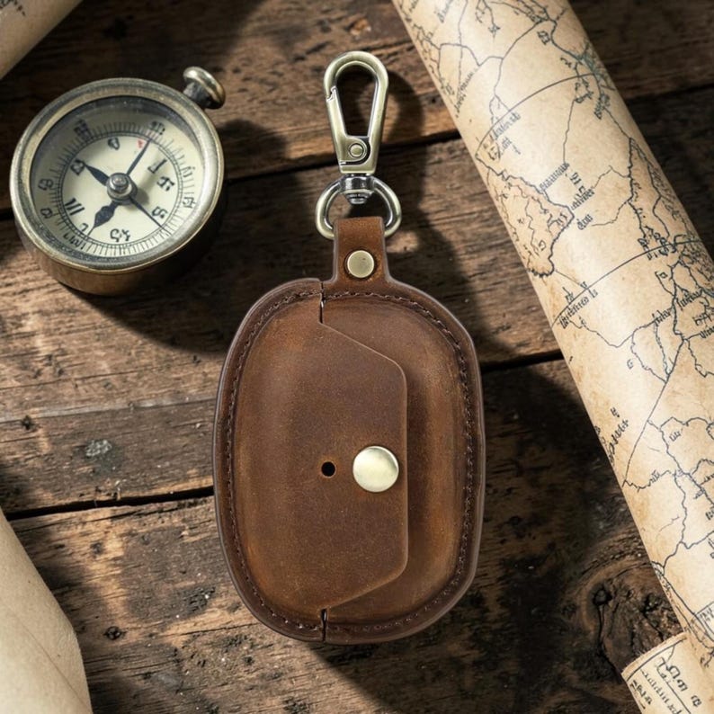 May include: A brown leather case with a brass clasp and button closure, designed to hold small electronics. The case is attached to a metal carabiner clip. A vintage compass and rolled map are in the background.