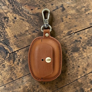 May include: A brown leather case with a brass clasp and a snap closure. The case is oval-shaped and has a stitched border. The clasp is attached to a metal ring. The case is designed to hold small items.