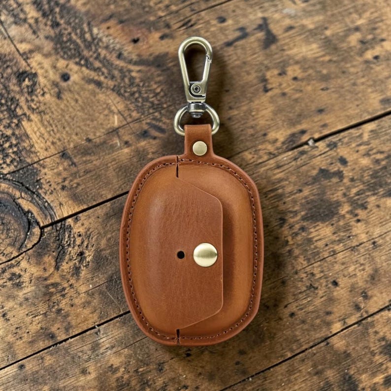 May include: A brown leather case with a brass clasp and a snap closure. The case is oval-shaped and has a stitched border. The clasp is attached to a metal ring. The case is designed to hold small items.