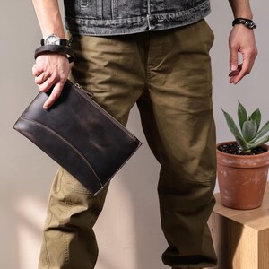 Puede incluir: Bolso de mano de cuero marrón oscuro con cierre de cremallera y costuras marrón claro. El bolso rectangular lo sostiene una persona que lleva un reloj y una pulsera. El fondo incluye una planta en maceta de terracota y una mesita de madera.