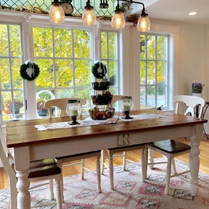 May include: A white farmhouse dining table with a wood top and turned legs. The table is set with a white tablecloth and a centerpiece of fruit and greenery. The table is surrounded by four white chairs with brown seats. The table is in a room with large windows and a chandelier with five light bulbs.