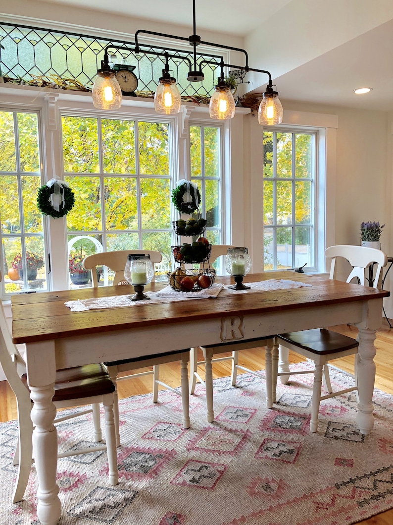 May include: A white farmhouse dining table with a wood top and turned legs. The table is set with a white tablecloth and a centerpiece of fruit and greenery. The table is surrounded by four white chairs with brown seats. The table is in a room with large windows and a chandelier with five light bulbs.