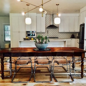 May include: A wooden dining table with eight clear plastic chairs. The table has a brown wood top and legs. The chairs are made of clear plastic and have a chrome frame. The table is set in a modern kitchen with white cabinets and a white tiled backsplash.