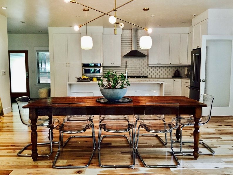 May include: A wooden dining table with eight clear plastic chairs. The table has a brown wood top and legs. The chairs are made of clear plastic and have a chrome frame. The table is set in a modern kitchen with white cabinets and a white tiled backsplash.