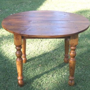 May include: A round wooden table with a natural wood finish. The table has a drop leaf design and turned legs.