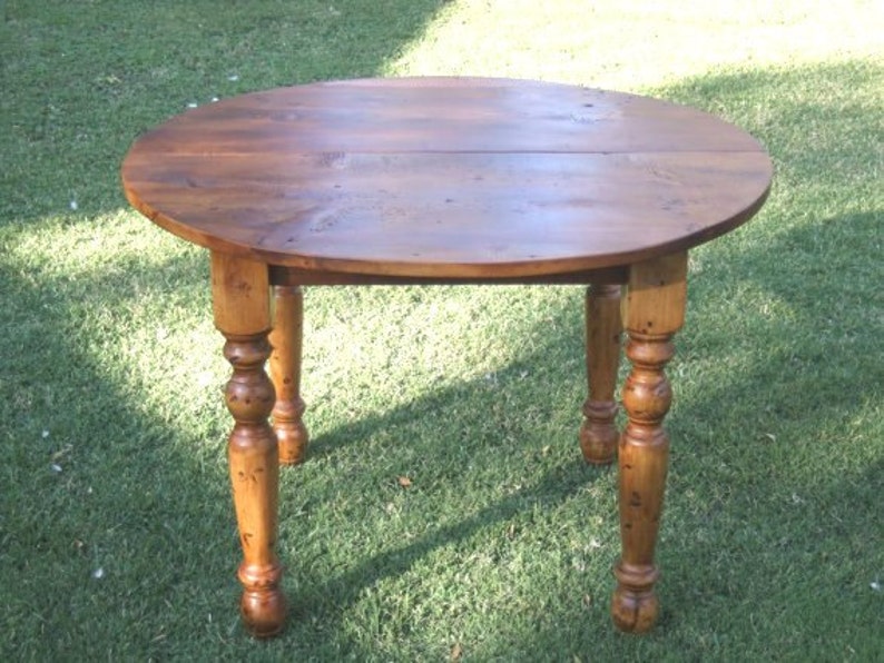 May include: A round wooden table with a natural wood finish. The table has a drop leaf design and turned legs.