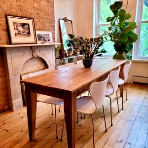 Pode incluir: Uma mesa de jantar de madeira r&uacute;stica com acabamento em madeira natural. A mesa &eacute; cercada por seis cadeiras brancas com pernas de metal dourado. A mesa est&aacute; localizada em uma sala com uma parede de tijolo, uma lareira e uma janela grande.