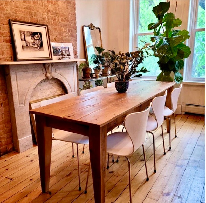 Pode incluir: Uma mesa de jantar de madeira r&uacute;stica com acabamento em madeira natural. A mesa &eacute; cercada por seis cadeiras brancas com pernas de metal dourado. A mesa est&aacute; localizada em uma sala com uma parede de tijolo, uma lareira e uma janela grande.