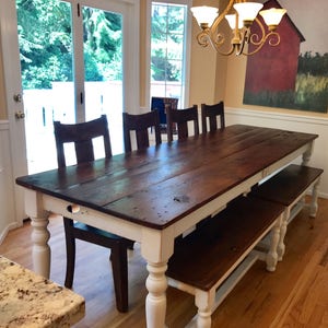 May include: A long, rustic farmhouse dining table with a white base and a dark wood top. The table is surrounded by chairs and a bench, all with a similar dark wood finish. The table is set in a dining room with a large window and a painting on the wall.