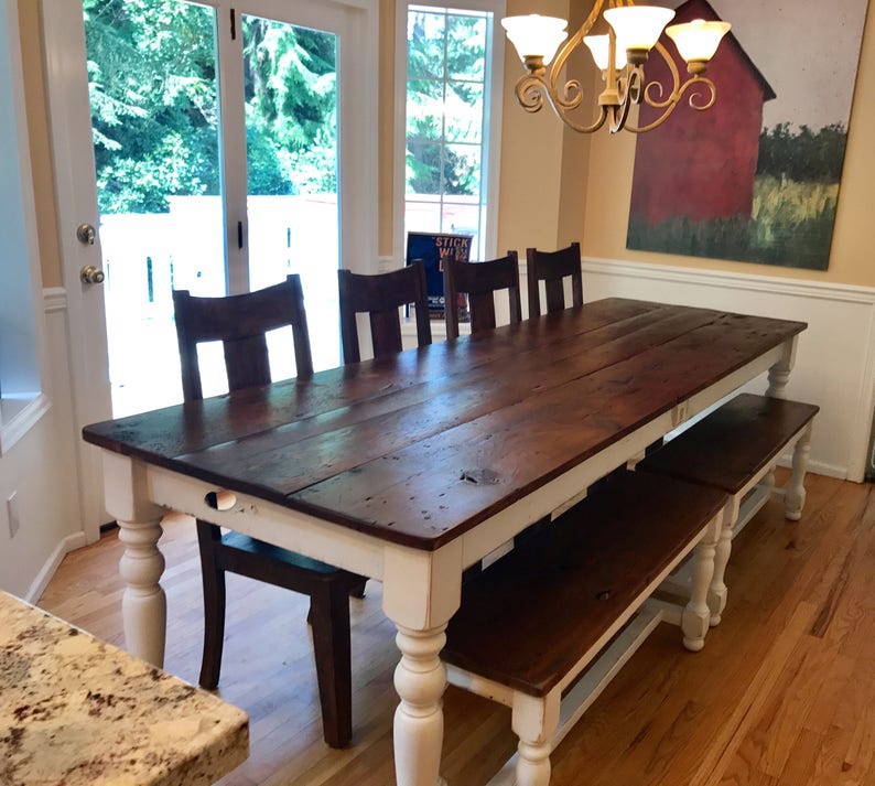 May include: A long, rustic farmhouse dining table with a white base and a dark wood top. The table is surrounded by chairs and a bench, all with a similar dark wood finish. The table is set in a dining room with a large window and a painting on the wall.