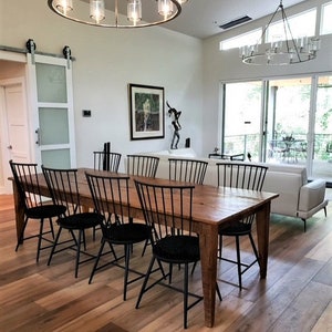 Pode incluir: Uma mesa de jantar de madeira comprida com cadeiras pretas. A mesa tem um acabamento r&uacute;stico e &eacute; cercada por cadeiras pretas com almofadas pretas. A mesa est&aacute; localizada em uma sala de jantar com pisos de madeira e uma grande janela.