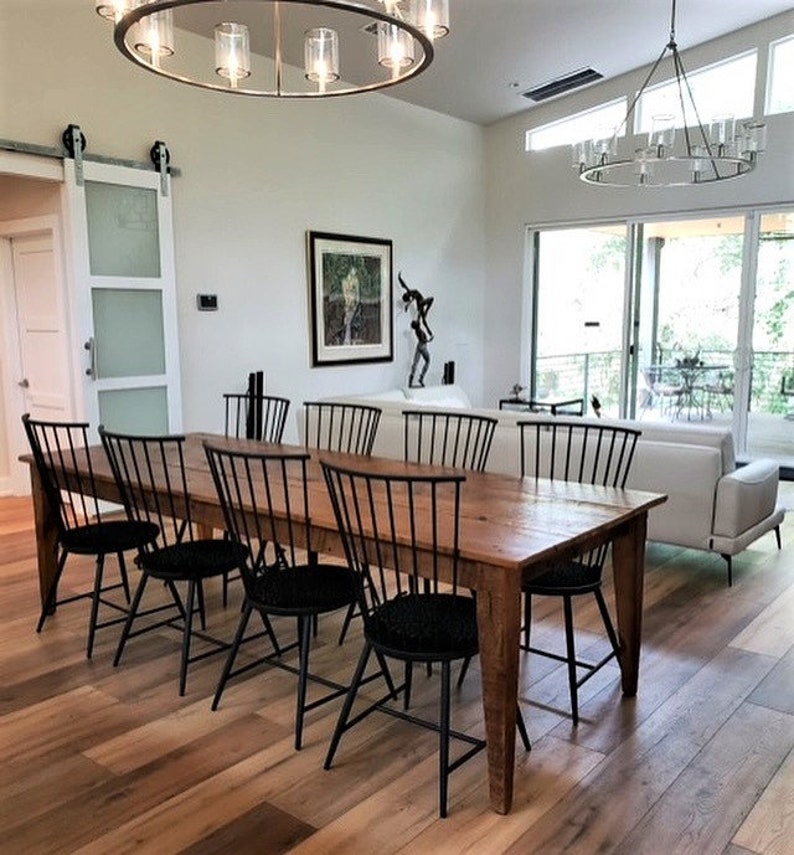 Pode incluir: Uma mesa de jantar de madeira comprida com cadeiras pretas. A mesa tem um acabamento r&uacute;stico e &eacute; cercada por cadeiras pretas com almofadas pretas. A mesa est&aacute; localizada em uma sala de jantar com pisos de madeira e uma grande janela.