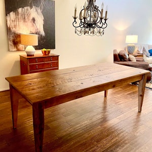 Pode incluir: Uma mesa de jantar de madeira r&uacute;stica com acabamento em madeira natural e um visual envelhecido. A mesa tem um tampo retangular e quatro pernas. A mesa est&aacute; em uma sala com piso de madeira e um lustre pendurado no teto.