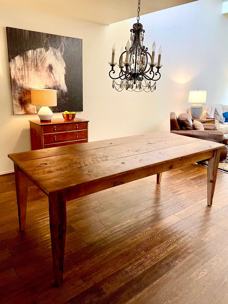 Pode incluir: Uma mesa de jantar de madeira r&uacute;stica com acabamento em madeira natural e um visual envelhecido. A mesa tem um tampo retangular e quatro pernas. A mesa est&aacute; em uma sala com piso de madeira e um lustre pendurado no teto.