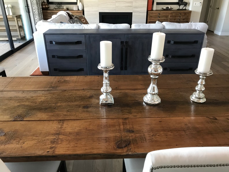 Pode incluir: Tr&ecirc;s casti&ccedil;ais de prata com velas brancas sobre uma mesa de madeira. A mesa &eacute; feita de t&aacute;buas de madeira recuperadas.