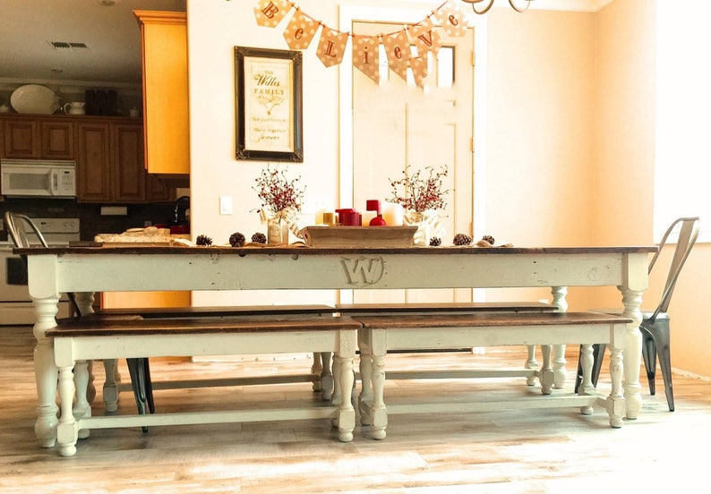 May include: A white farmhouse style dining table with two matching benches. The table has a distressed white finish and a dark wood top. The table is decorated with a red and gold centerpiece and a banner that reads "Believe".