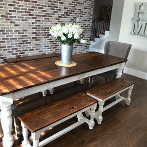May include: A farmhouse style dining table with a white base and a brown wood top. The table is surrounded by two matching benches with white bases and brown wood tops. The table is set against a brick wall.