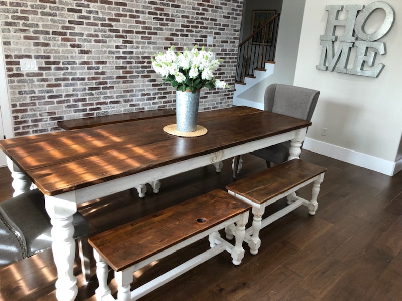 May include: A farmhouse style dining table with a white base and a brown wood top. The table is surrounded by two matching benches with white bases and brown wood tops. The table is set against a brick wall.