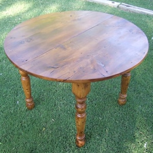 May include: A round wooden table with a natural wood finish and turned legs. The table is made of solid wood and has a rustic look.
