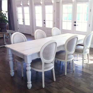 May include: A white farmhouse table with six white chairs. The table has a distressed white finish and turned legs. The chairs have a linen fabric seat and back.
