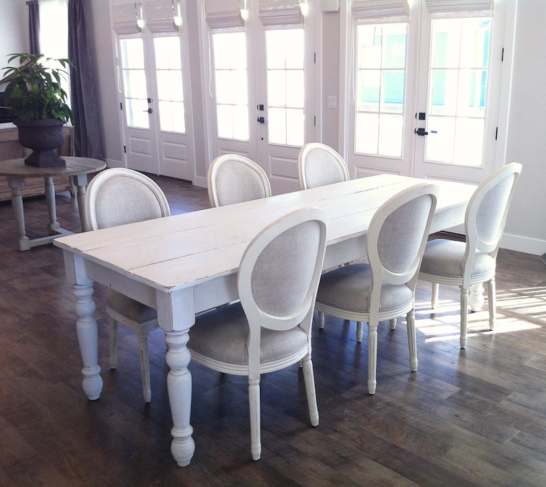 May include: A white farmhouse table with six white chairs. The table has a distressed white finish and turned legs. The chairs have a linen fabric seat and back.
