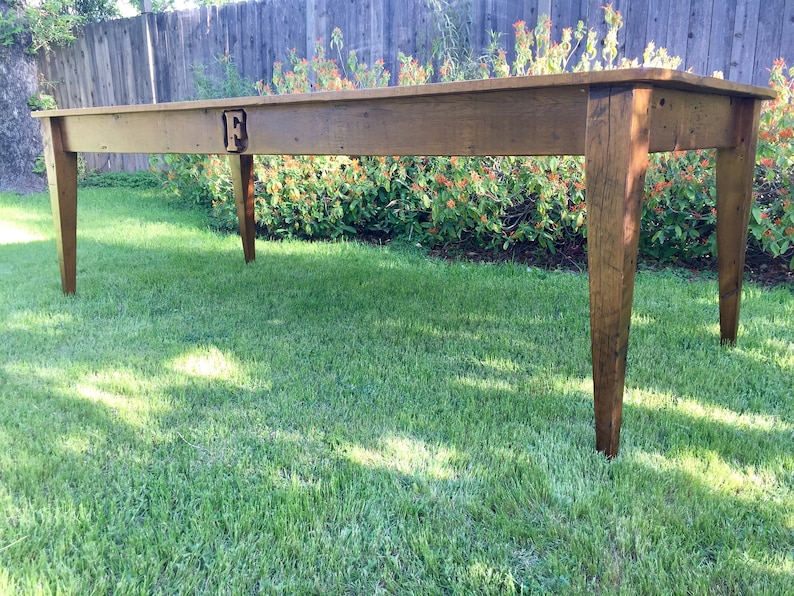 Pode incluir: Uma mesa longa de madeira com um design simples. A mesa tem um acabamento de madeira natural e &eacute; feita de madeira maci&ccedil;a. A mesa tem uma &uacute;nica letra grande "F" esculpida na superf&iacute;cie superior. A mesa est&aacute; sentada em um gramado.