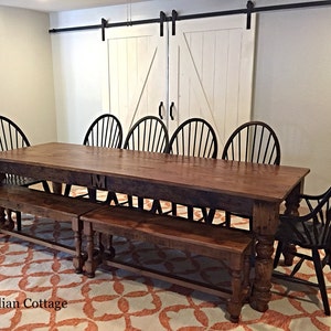 May include: A large wooden dining table with two matching benches. The table has a dark wood finish and is made of reclaimed wood. The table is set with black chairs and has a single initial "M" carved into the top. The table is in a room with white painted sliding barn doors.