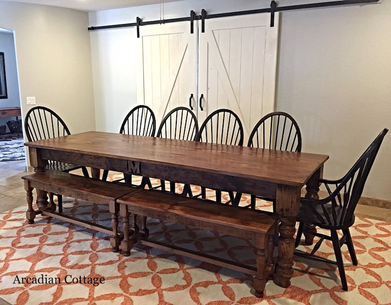 May include: A large wooden dining table with two matching benches. The table has a dark wood finish and is made of reclaimed wood. The table is set with black chairs and has a single initial "M" carved into the top. The table is in a room with white painted sliding barn doors.