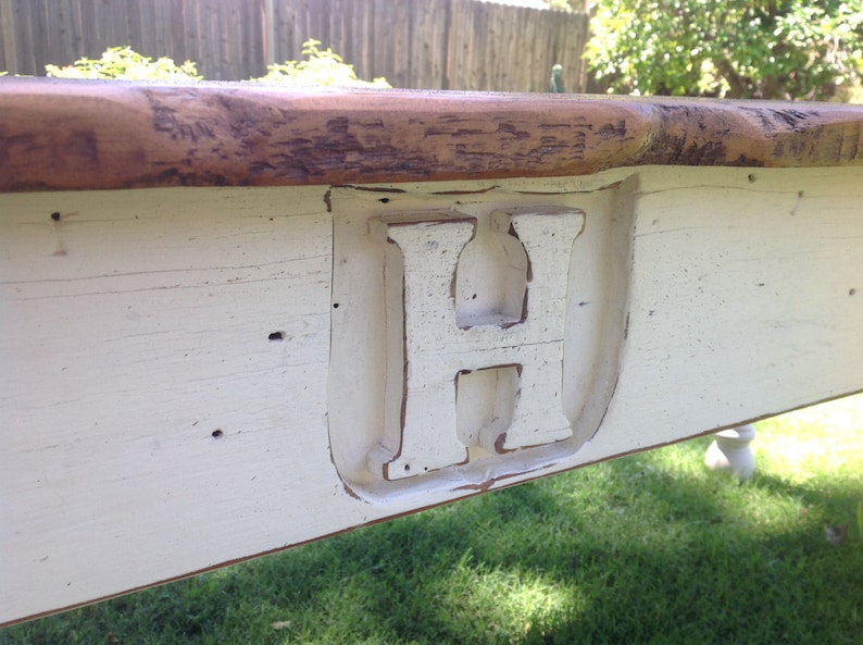May include: Close-up of a white painted wooden table top with a large, white painted letter "H" carved into the surface. The table top has a distressed, rustic look.