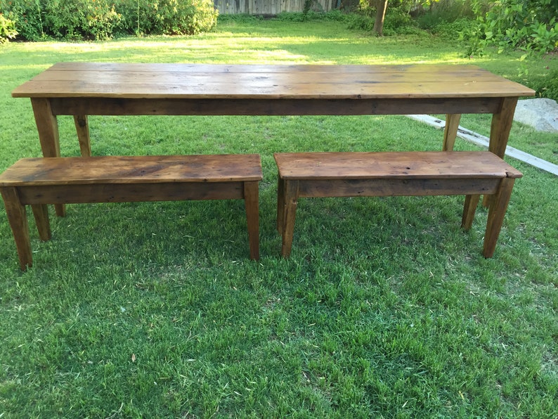 Pode incluir: Uma mesa de madeira r&uacute;stica e dois bancos colocados em um gramado verde. A mesa &eacute; longa e estreita com um acabamento de madeira natural. Os bancos s&atilde;o mais curtos e largos, tamb&eacute;m com um acabamento de madeira natural.