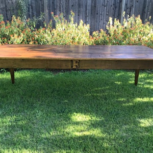 Pode incluir: Uma mesa de madeira r&uacute;stica longa com um acabamento marrom escuro. A mesa tem um design simples com quatro pernas e uma superf&iacute;cie retangular. A mesa est&aacute; sentada em um gramado verde.