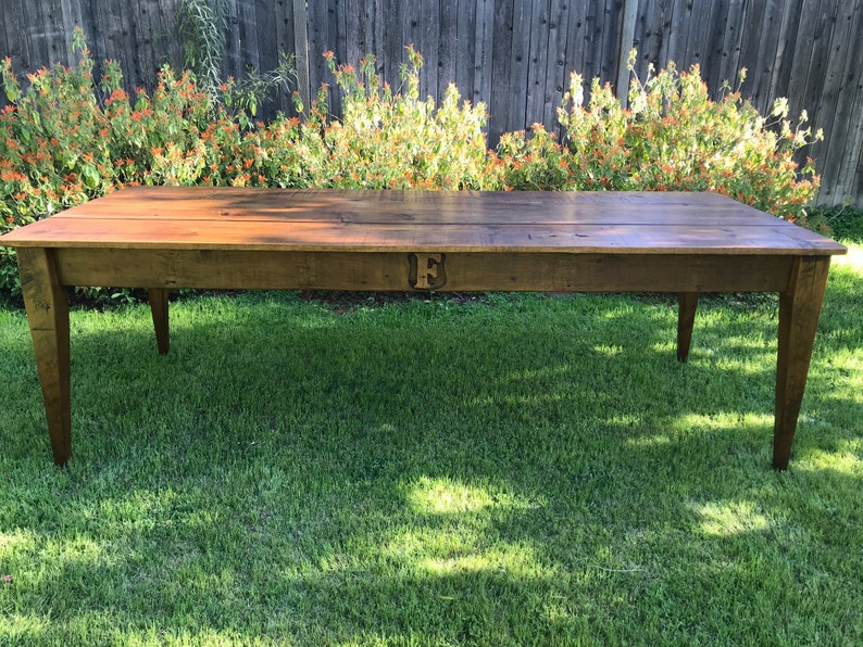 Pode incluir: Uma mesa de madeira r&uacute;stica longa com um acabamento marrom escuro. A mesa tem um design simples com quatro pernas e uma superf&iacute;cie retangular. A mesa est&aacute; sentada em um gramado verde.