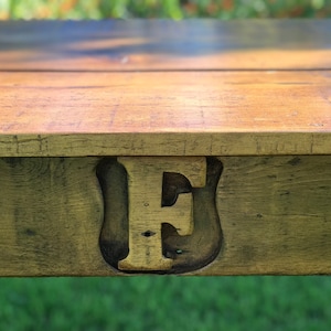 Pode incluir: Close-up de uma mesa de madeira com uma grande letra "E" pintada de ouro esculpida na superf&iacute;cie. A madeira tem um aspecto r&uacute;stico e desgastado.