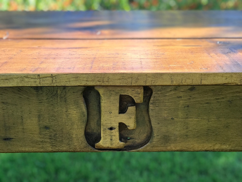 Pode incluir: Close-up de uma mesa de madeira com uma grande letra "E" pintada de ouro esculpida na superf&iacute;cie. A madeira tem um aspecto r&uacute;stico e desgastado.