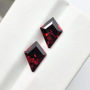 Könnte beinhalten: Zwei lose facettierte Edelsteine in einer tiefroten Farbe und einer ausgeprägten Drachenform. Jeder Stein hat eine spitze Ober- und Unterseite mit abgewinkelten Kanten und einen Brillantschliff, der das Licht auf den dunkelroten Oberflächen reflektiert.