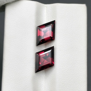 Peut inclure: Deux pierres précieuses rouge foncé à facettes en forme de losange. Les gemmes ont un centre carré et des côtés inclinés, reflétant la lumière. Les gemmes sont posées sur un fond blanc.