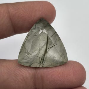 May include: A faceted, triangular-shaped rutilated quartz gemstone featuring a translucent, pale green body with distinct, dark needle-like inclusions scattered throughout the stone, creating a unique pattern within the clear crystalline structure.